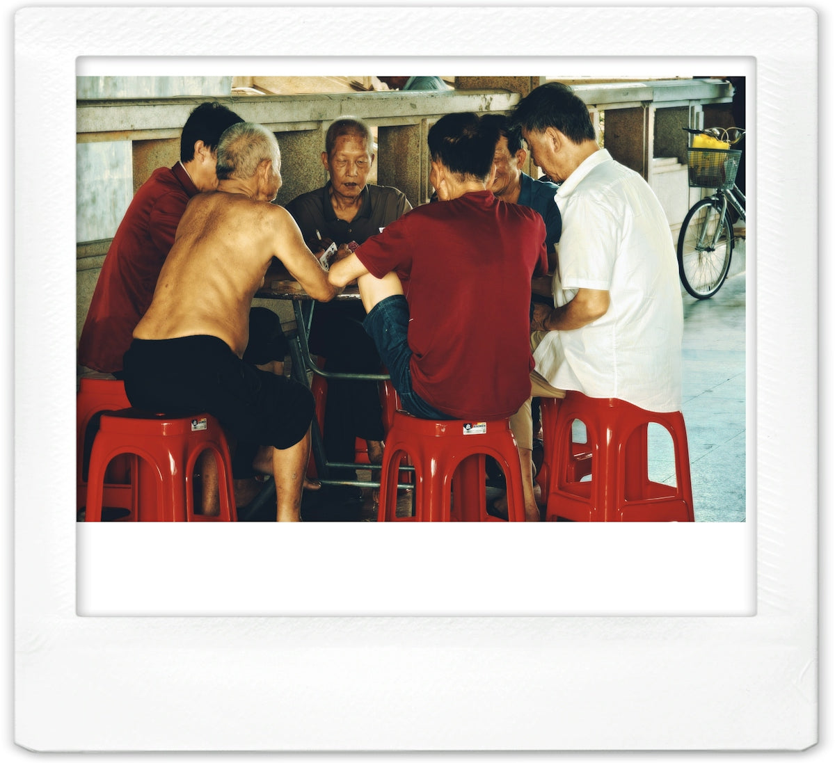 A group of men playing cards on red plastic stools in a covered area